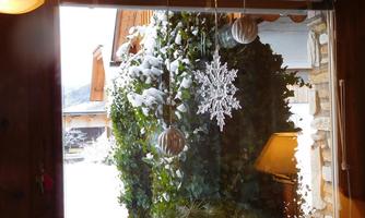 A wintry view through a window, with snow and a carved snowflake in the foreground. Outside, green plants and a lit lamp are visible.
