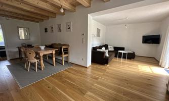 A bright and modern living room with wooden ceilings and a cozy dining area. The decor is simple, with a sofa, a table, and a television.