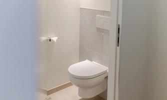 A simple, modern bathroom with a toilet and bright, neutral design. The walls are light and the floor is tiled beige.