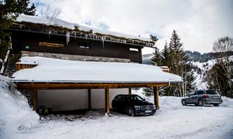 A wooden house in the snow with a covered parking space. Two cars are parked in front of the house, surrounded by snow-covered trees.