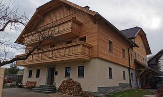A modern designed wooden house with two floors and balconies. In the foreground, you can see wood stacks and a well-maintained garden area.