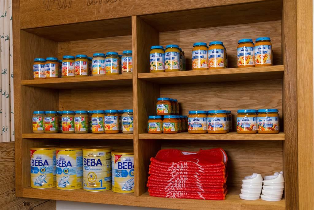 A wooden shelf with several jars of baby food and cans of formula. Below, there is a stack of red towels and white plates.