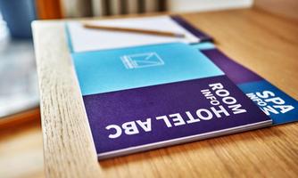 A stack of informational brochures about the hotel, including room service and spa details, is on a wooden shelf. Next to the brochures is a ballpoint pen and a notepad.