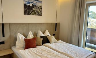 A cozy bedroom with two separate beds and decorative pillows. A picture of mountains hangs on the wall, and there is a balcony with a view outside.