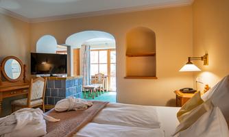 A cozy hotel room with a double bed and soft colors. In the background, there is a television and a seating area with a table and chairs.