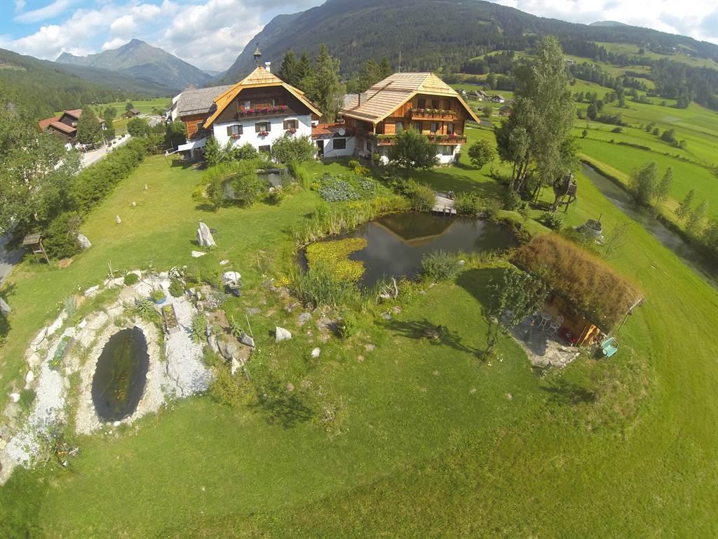 A picturesque farmhouse surrounded by green meadows and mountains. In the foreground, there is a small pond and beautifully arranged gardens.