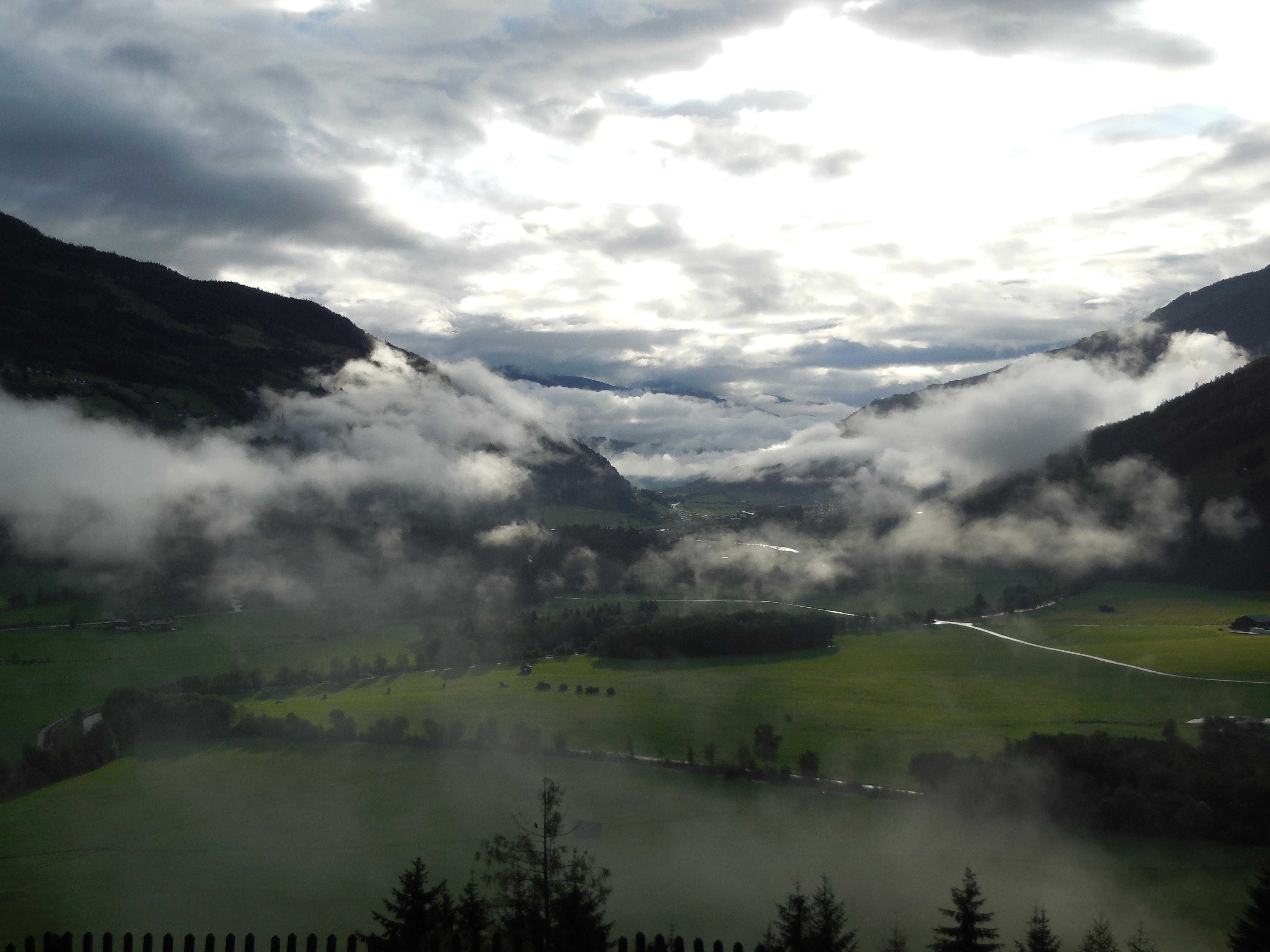 A picturesque view of a valley with green meadows and gentle hills. Mist floats through the landscape, giving the scene a mystical ambiance.