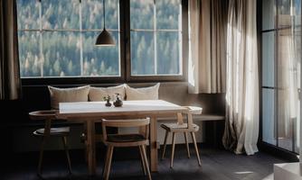 A modern furnished room with a wooden dining table and chairs. Large windows let in plenty of daylight and offer a view of nature.