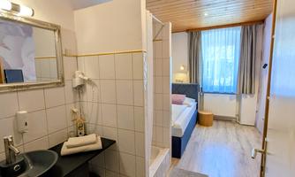 A bedroom with an integrated bathroom area and shower. The room is bright and cheerful, with a wooden ceiling and large windows.