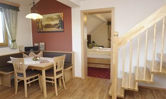 A cozy apartment with a dining area and a wooden staircase. In the background, there is a bedroom with a bed.