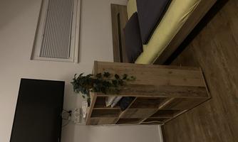 A simple bedroom with a bed, a wooden box, and a television on the wall. The walls are light, and the room has a cozy wooden floor.