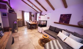 A modern living room with a cozy sofa, a dining table, and wooden ceiling beams. The room is bright and elegantly furnished, with an open kitchenette.