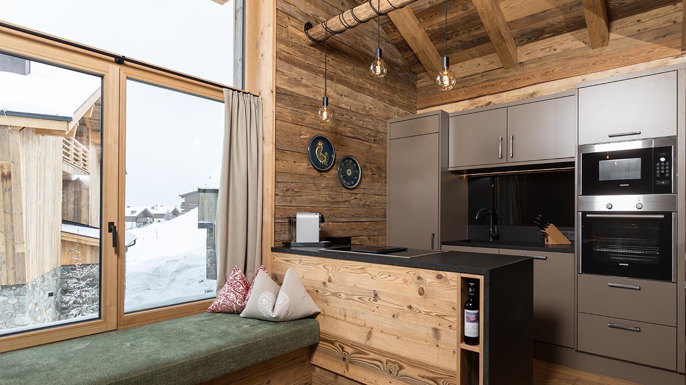 A modern kitchen with wooden details and large windows. The view shows a snow-covered landscape.