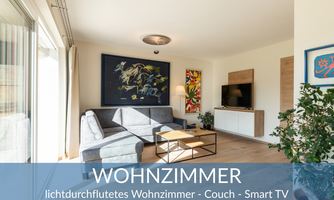 Bright living room with a gray couch and a smart TV. Modern artworks adorn the walls, and large windows provide plenty of daylight.