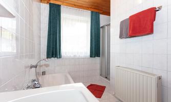 A bright bathroom with a bathtub and a shower. Red towels hang on the wall, and the windows are fitted with green curtains for privacy.
