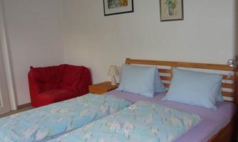 A simple bedroom with a double bed and blue pillows. Next to the bed is a red armchair and a small lamp.