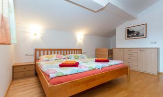 A cozy bedroom with a double bed and colorful bedding. The room has a bright wooden floor and a modern dresser.