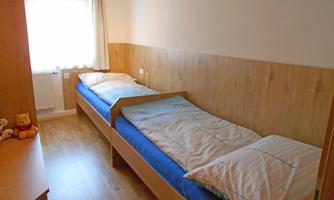 A simple bedroom with two single beds and wooden paneling. The window provides natural light, and there are a few personal items in the room.