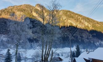 A winter landscape with snow-covered mountains and trees. In the foreground, simple wooden houses and a fence are visible.