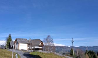 An old, charming house stands in a rural setting. In the background, mountains and a clear blue sky can be seen.