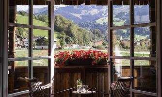 View from an open window of a picturesque landscape with green hills and mountains in the background. On the balcony, there are two chairs and a blooming flower bench.