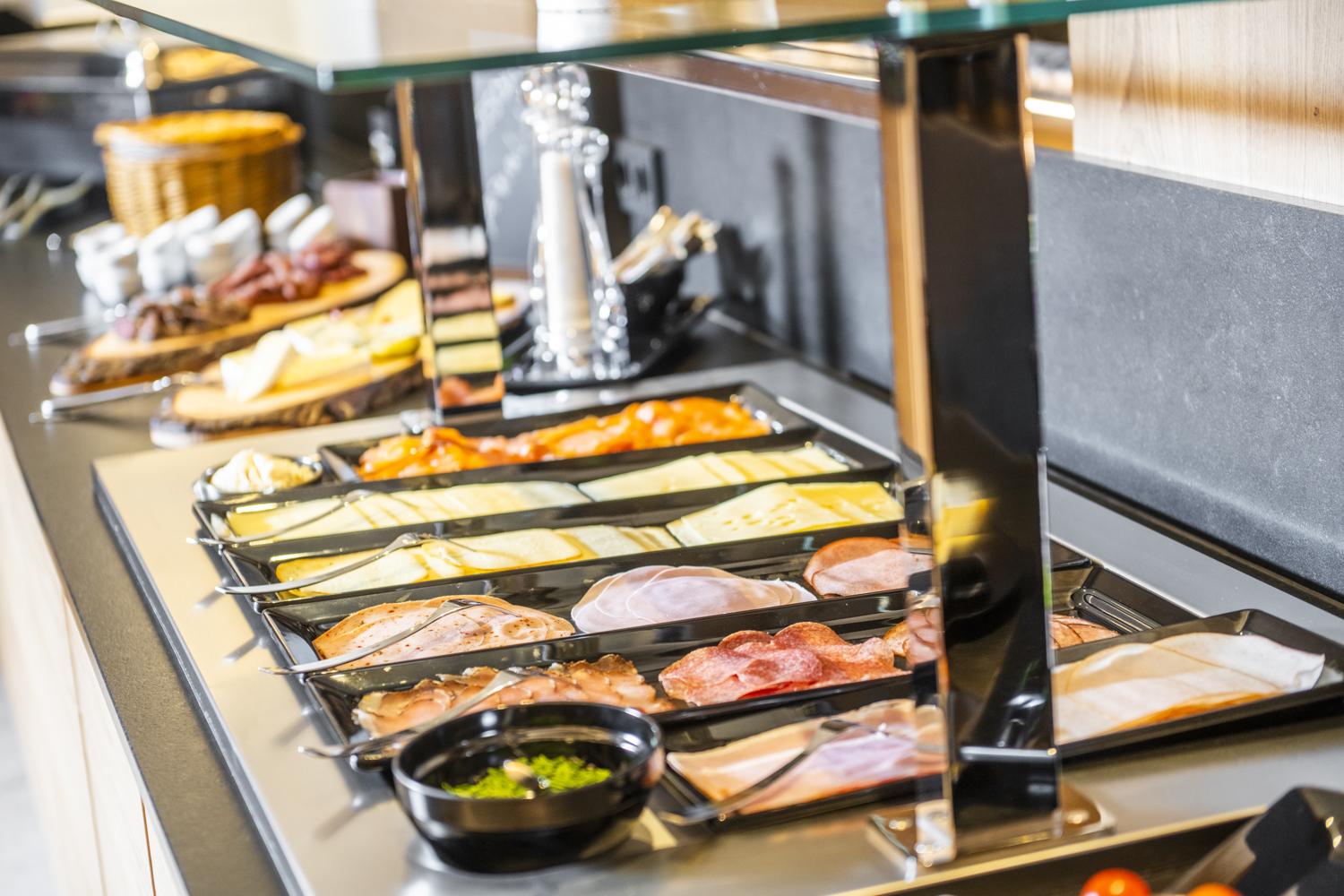 A buffet with various types of cold cuts and cheeses on a table. Next to it is a bowl of olives.