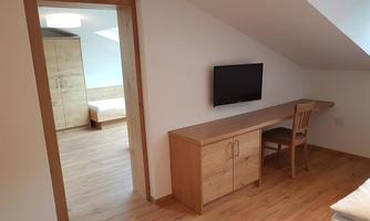A bright, modern room with wooden furniture and a TV. The room features a large window front and a desk with a chair.