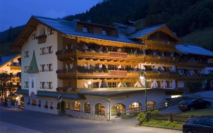 A pretty hotel with wooden decorations that is illuminated at night. The surroundings are surrounded by mountains and convey a cozy atmosphere.