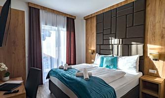 A modern hotel room with a double bed and wooden furniture. The walls are painted in warm tones and there are large windows with curtains.