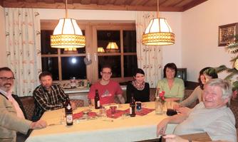 A group of seven people is sitting at a table in a cozy room. There are drinks and an inviting atmosphere with warm light.