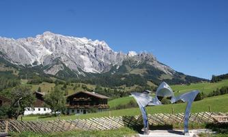 A modern sculpture stands in a picturesque mountain landscape. In the background, impressive mountains and traditional buildings can be seen.