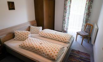 A cozy bedroom with a double bed and two pillows. The room has a wardrobe and a chair next to a window with curtains.