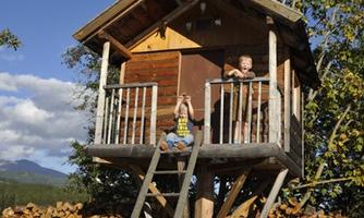 A treehouse with wooden log walls and a sunny environment. Two children are standing on the platform and enjoying the view.
