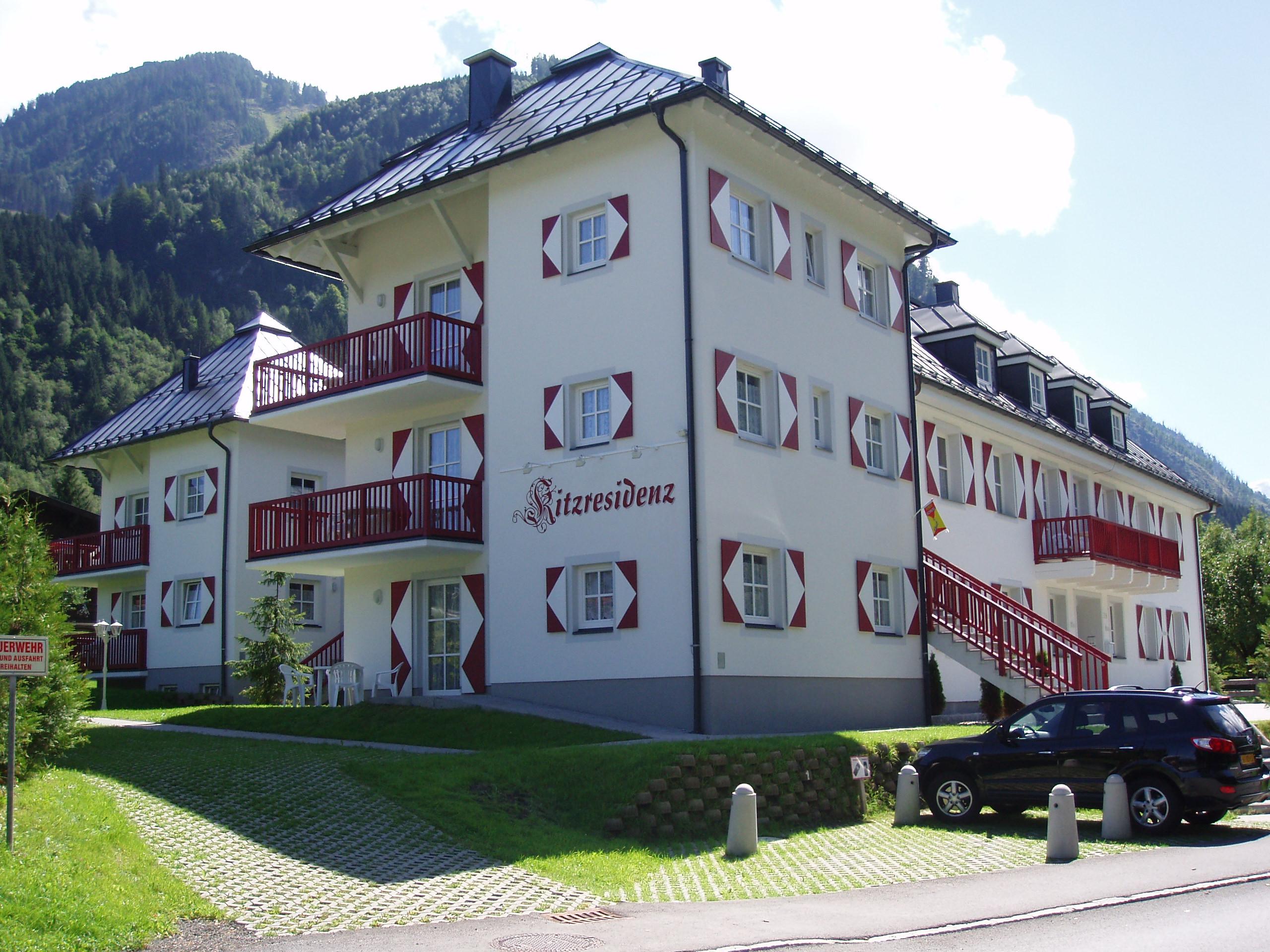 A modern building with several floors and balconies, surrounded by green landscape. The facade is bright with red and white accents.