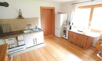 A bright kitchen with wooden flooring and large windows. There is a sink, a refrigerator, and a wooden cupboard.