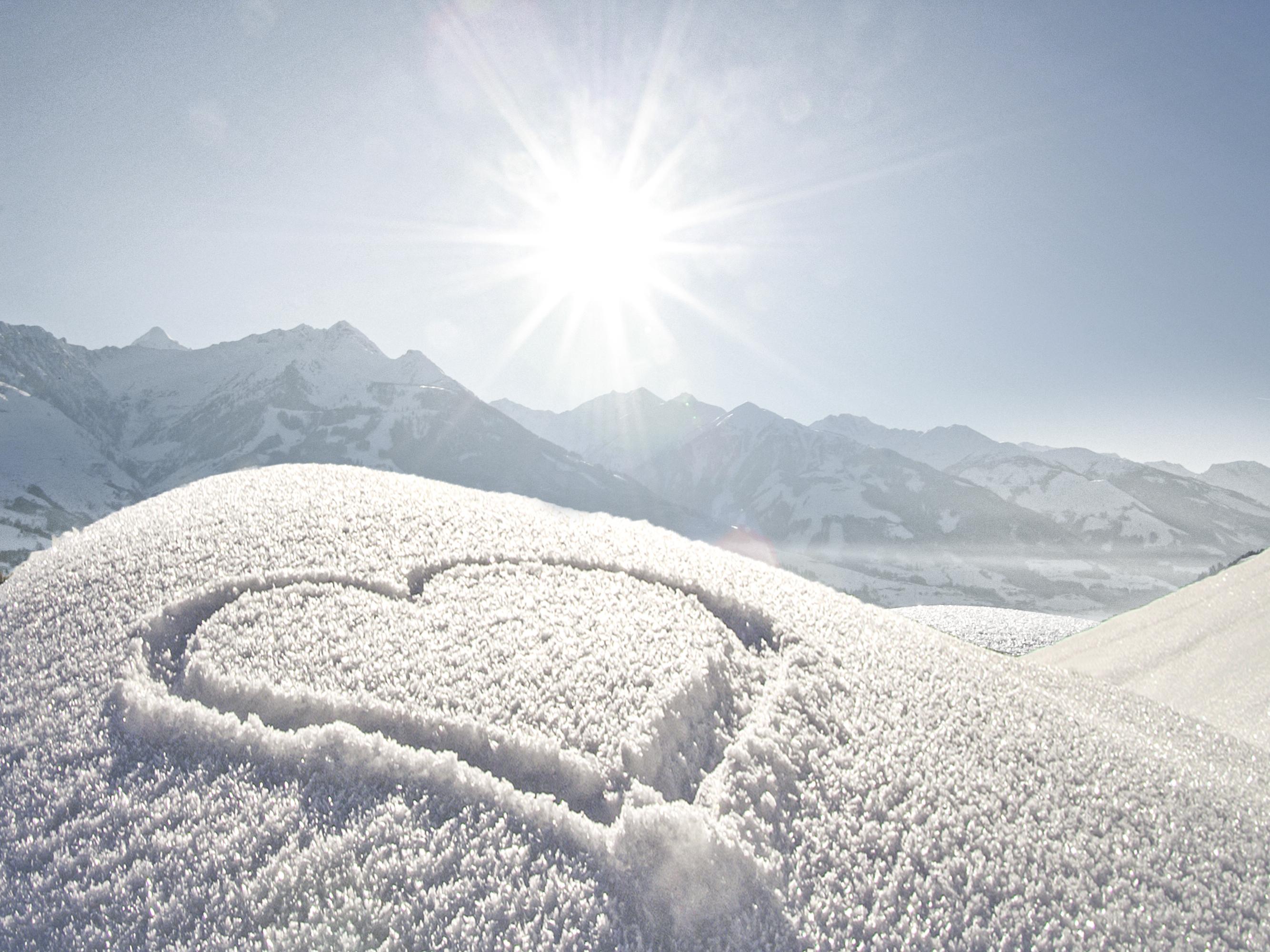 A snowy landscape with a heart scratched into the snow. The sun is shining brightly over the mountains.