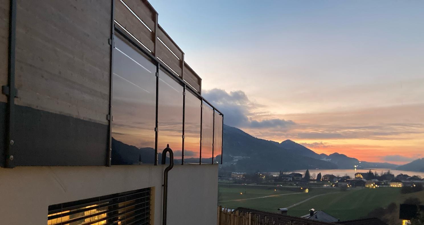 A modern building with large windows and a view of a picturesque landscape. In the background, a colorful sunset illuminates the sky above the green fields and mountains.