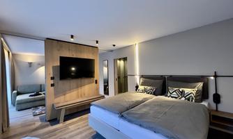 A modern hotel room with a double bed and a television on the wall. In the background, there is a cozy sitting area with a sofa.
