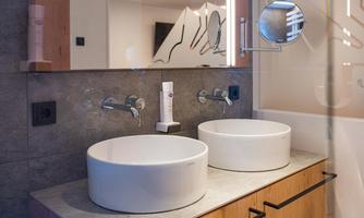 A modern bathroom with two round sinks and a wooden drawer. The wall is in neutral colors, complemented by stylish mirrors and fixtures.