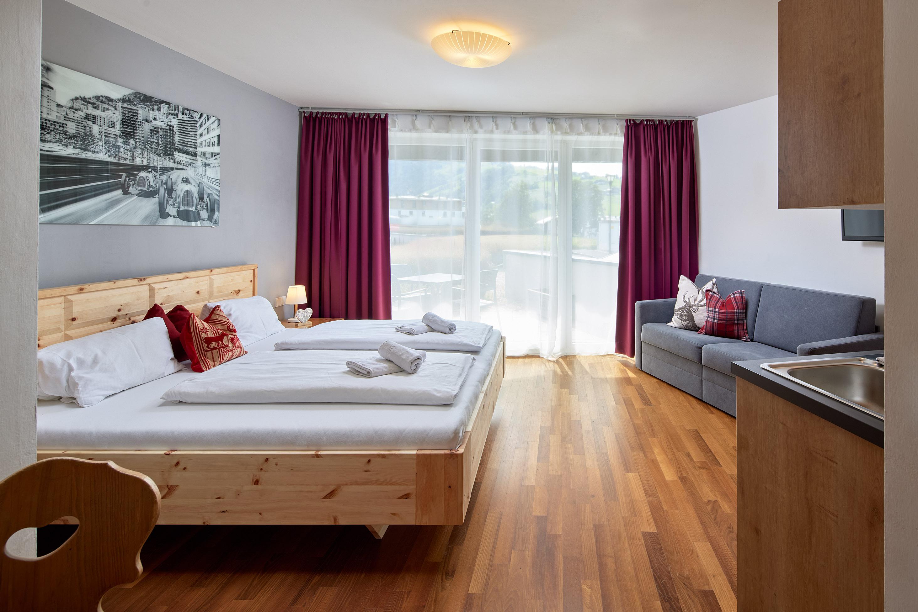 A modern room with two beds, a sofa, and a small kitchenette. Large windows provide plenty of daylight and a friendly atmosphere.