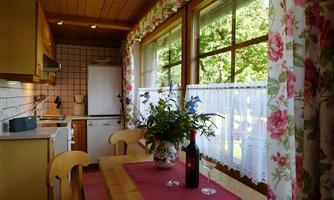 A cozy kitchen with wooden surfaces and large windows. On the table is a vase with flowers and a bottle of wine.