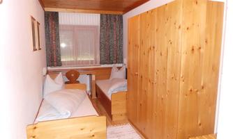 A simple bedroom with two wooden single beds. The room has wooden-clad furniture and windows with curtains.