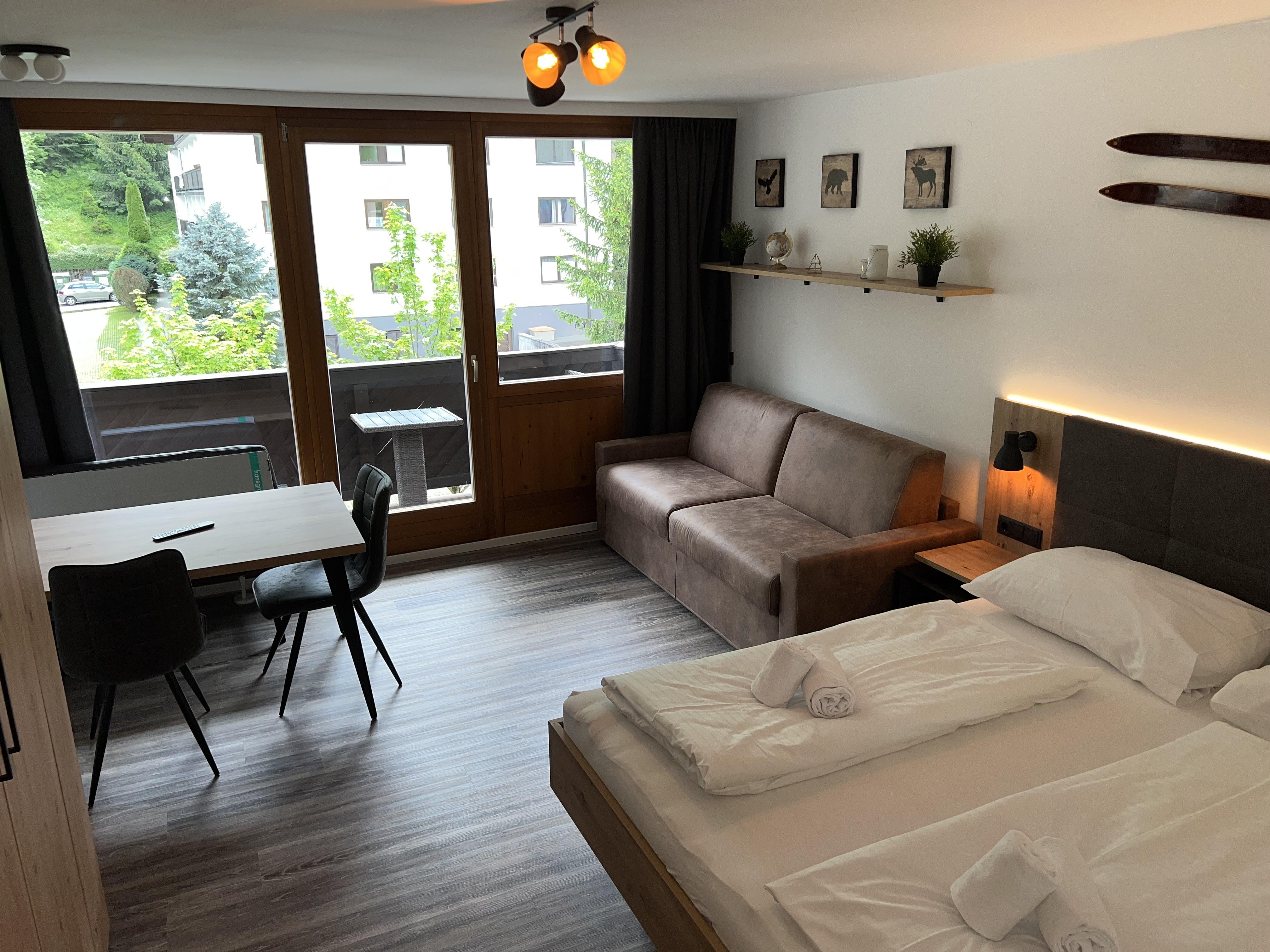 A modern room with a double bed, sofa, and table. Large windows provide light and a view of the outdoors.