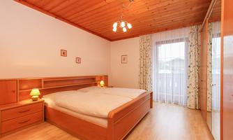 A cozy bedroom with a wooden bed and bright walls. Large windows let in plenty of daylight.