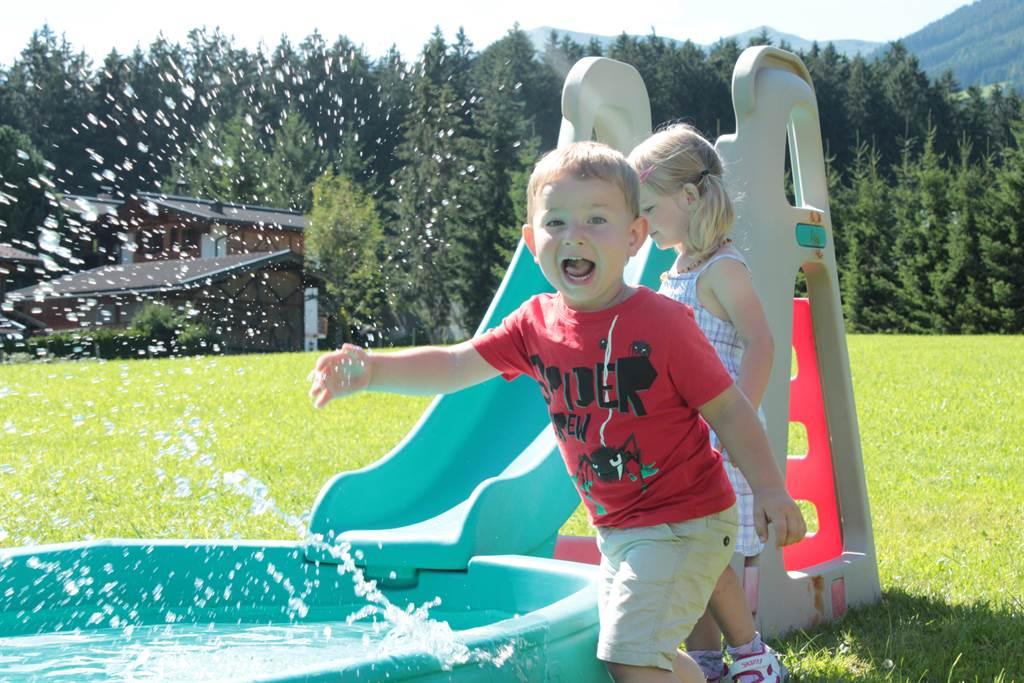 A boy happily splashes in the water, standing next to a slide. In the background, trees and a house can be seen.