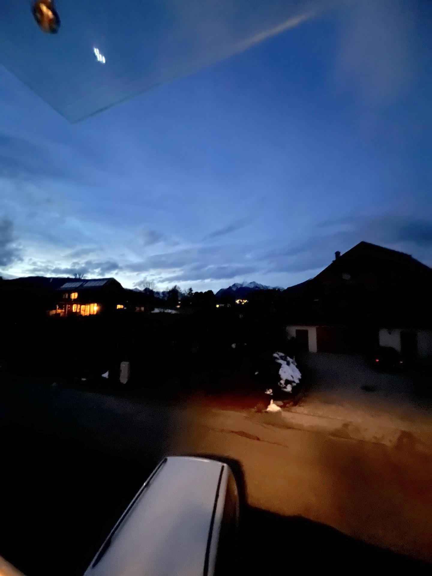 A peaceful evening with a dark sky and soft clouds. In the background, some illuminated houses and snow-covered mountains can be seen.