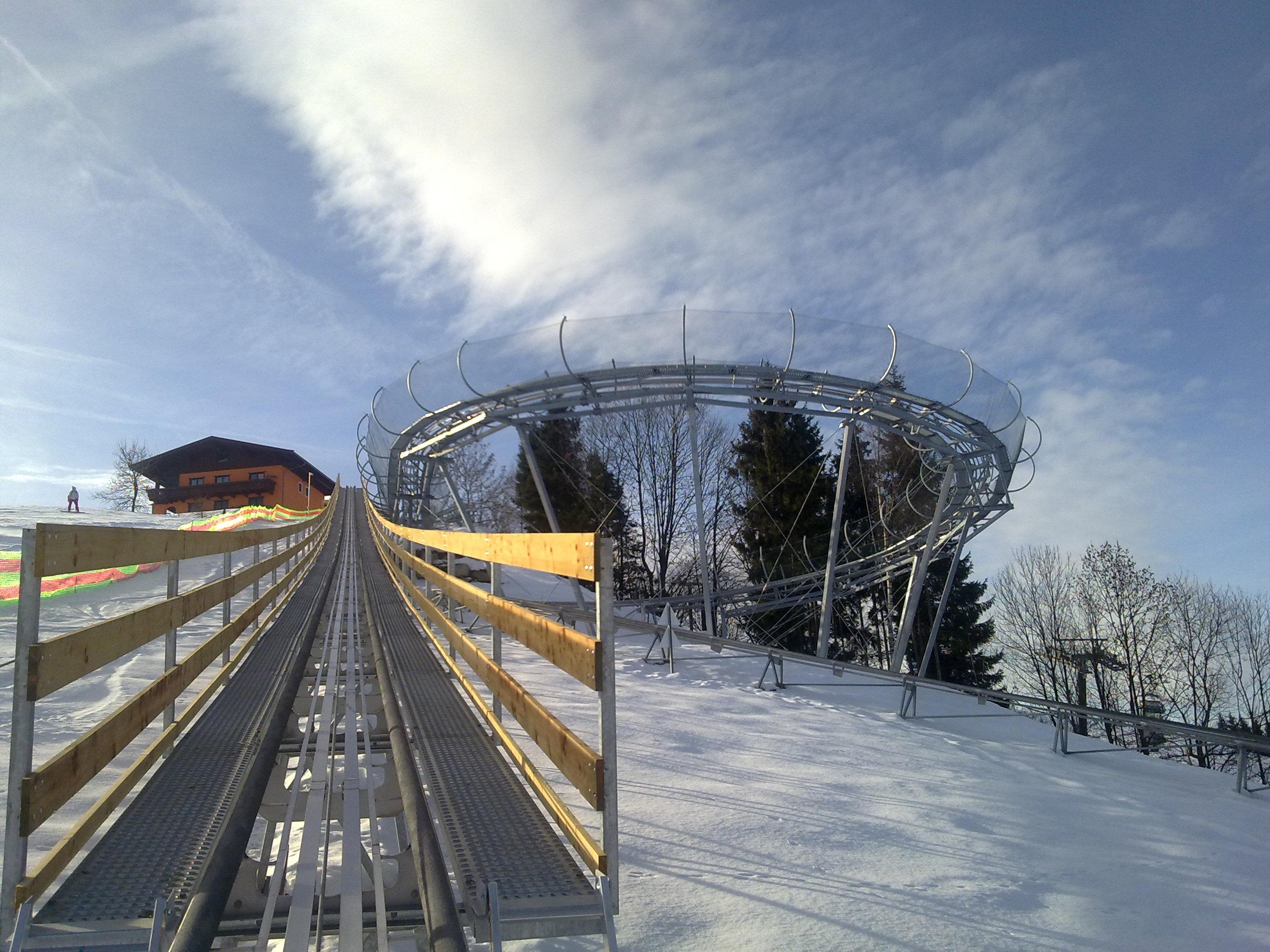A toboggan run that goes through snow, with a winding track in the background. The sky is clear and the surroundings are wintry.