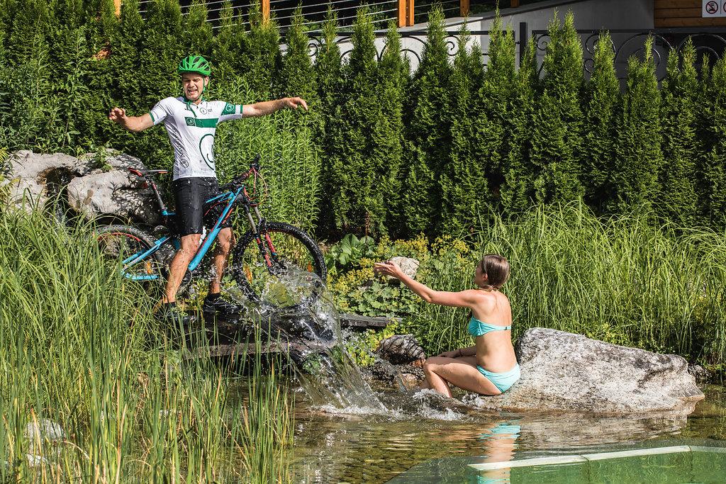 A man on a mountain bike balances on a rock next to a waterfall. A woman sits in the water and watches him.