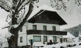 A snow-covered house in winter with a large tree in the foreground. The surroundings are quiet and idyllic.