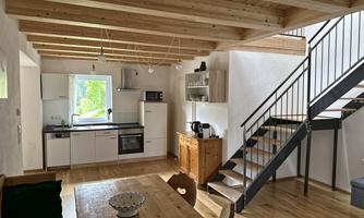 A modern and bright kitchen with wooden shelves and a dining table. A staircase leads to the upper area of the room.